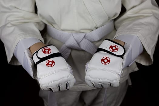 Young woman in kimono and white belt shows clenched fists in protective gloves in Kyokushin karate, combat equipment and protection of a martial arts athlete