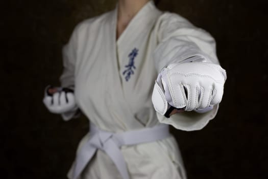 Girl athlete in kimono and white belt in Kyokushin karate hits a blow in protective gloves with clenched fist. Strong blow in martial arts. Inscription in Japanese: Kyokushinkai