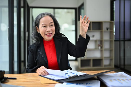 Senior businesswoman working at desk and waving hand greeting someone with smiling cheerfully.
