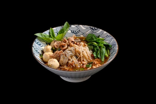 A bowl of Spicy Thai noodles soup with pork offal, meatballs and vegetables on black background.