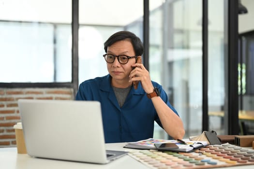 Professional senior male interior designer having phone conversation with client and using laptop at desk.