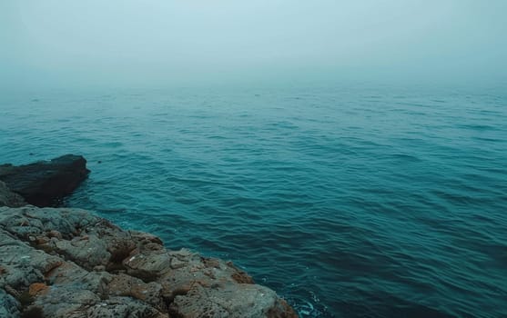 A hazy seascape where the calm ocean meets a rocky shoreline under a foggy sky. The subdued colors paint a picture of stillness and mystery