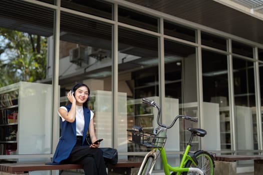 Businesswoman listening to music with headphones while sitting by her bicycle outside office. Concept of modern technology, relaxation, and sustainable commuting.