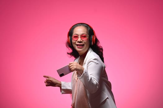 Happy senior woman woman listening to music and dancing on light pink background.