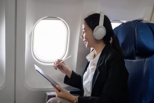 Businesswoman with tablet and headphones looking out airplane window. Concept of in flight productivity and remote work.