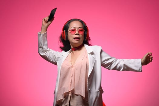 Portrait of charming retired woman listening music with headphones dancing on light pink background.