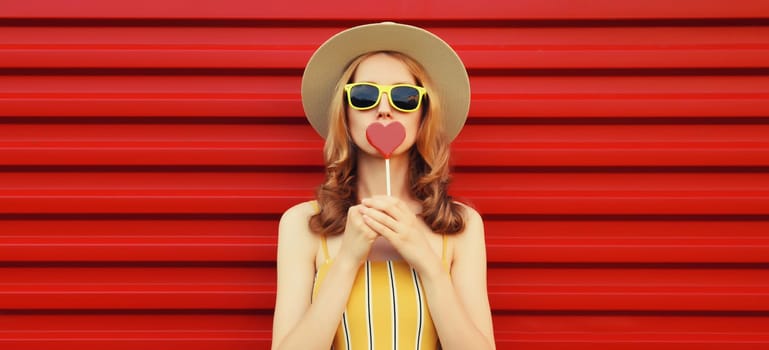 Summer portrait of beautiful young woman with sweet lollipop blowing kiss wearing yellow sunglasses, tourist straw round hat on bright red background