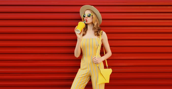 Beautiful stylish happy young woman drinking fresh juice holding cup wearing summer hat, yellow outfit posing on red wall background