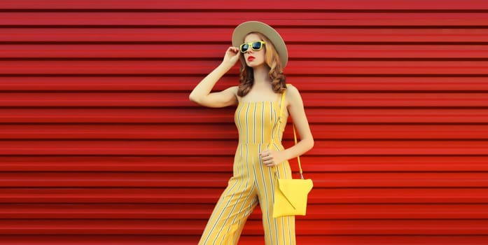 Beautiful trendy young woman model posing with handbag wearing yellow jumpsuit, summer straw hat on red background