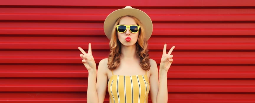 Portrait of beautiful stylish young woman blowing kiss in summer hat, yellow sunglasses posing on red wall background