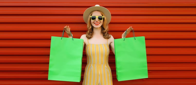 Stylish beautiful happy smiling young woman posing with bright green shopping bags wearing summer straw hat on red wall background