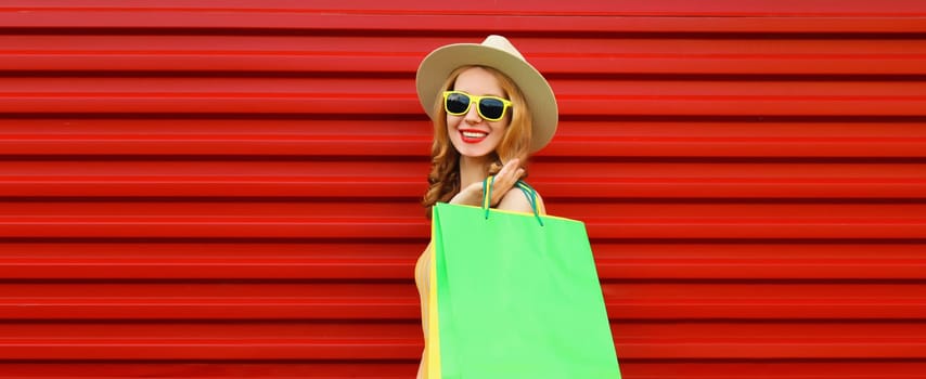 Stylish beautiful happy smiling young woman posing with bright green shopping bags wearing summer straw hat on red wall background