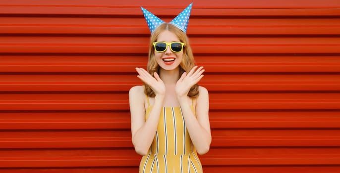 Holiday, celebration, event. Happy cheerful young woman laughs celebrating having fun at a party wearing festive birthday hat on red background