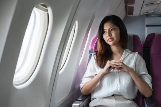 Asian woman sitting in airplane looking out window with thoughtful expression. Concept of air travel, journey, and relaxation.