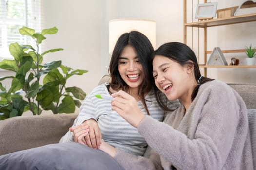 Asian lesbian couple celebrating positive pregnancy test at home. Concept of love, family planning, and LGBTQIA parenthood.