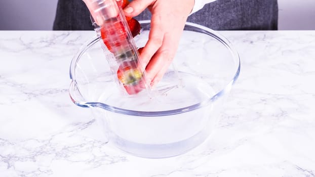 Ripe strawberries are submerged in water within a large glass mixing bowl, a step in washing the fruit to ensure cleanliness and longevity before storage or consumption.