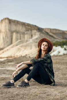Serene young woman in hat sitting peacefully in front of majestic mountain landscape on sunny day