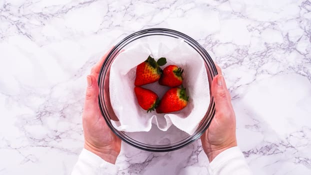 Flat lay. Strawberries, freshly washed and dried, are neatly stored in a glass bowl lined with a paper towel and securely covered with plastic wrap to maintain freshness.