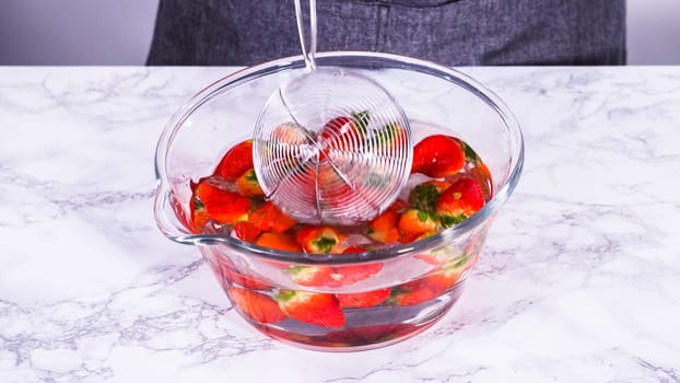 Ripe strawberries are submerged in water within a large glass mixing bowl, a step in washing the fruit to ensure cleanliness and longevity before storage or consumption.