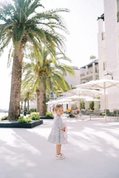 Little girl with a pink toy rabbit stands on a street with green palm trees. Side view. High quality photo