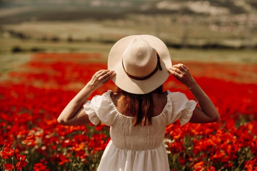 Field poppies woman. Happy woman in a white dress and hat stand through a blooming field of poppy raised her hands up. Field of blooming poppies