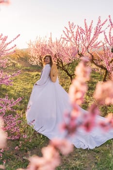 Woman blooming peach orchard. Against the backdrop of a picturesque peach orchard, a woman in a long white dress and hat enjoys a peaceful walk in the park, surrounded by the beauty of nature