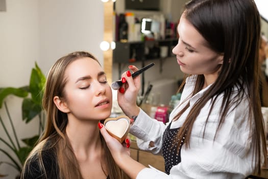 Young beautiful makeup artist lightly applies highlighter to the clients face at beauty salon