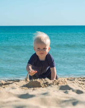A joyful blonde baby boy is crawling with sand at the beach on a sunny day. The clear blue ocean waves are calmly visible in the background, providing a picturesque and serene setting.