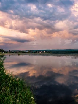 A tranquil river reflects the colorful sunset sky with clouds in a rural countryside setting. The serene scene is surrounded by lush greenery, creating a picturesque and peaceful environment.