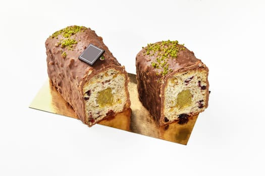 Sliced mini fruitcake with dried berries, milk chocolate glaze and pistachio sprinkles, displayed on golden cardboard isolated on white. Sweet pastries concept