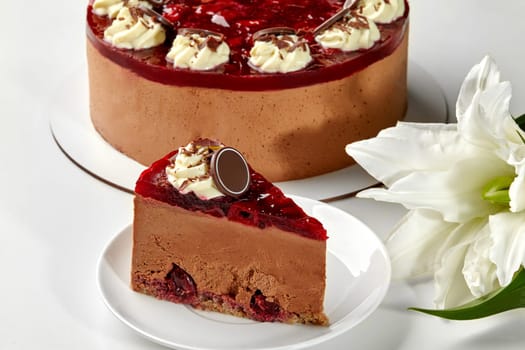 Slice of rich chocolate cheesecake with moist sponge and sweet cherries topped with berry jelly, whipped cream, and chocolate shavings, on background of whole cake and white flowers