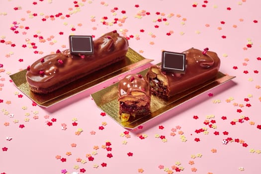 Appetizing milk chocolate bars on shortcrust cookies with fruit jelly and pecans decorated with dried berry crumbs on golden cardboard, against pink background with colorful flower shaped confetti