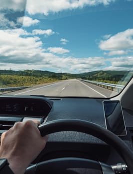 A driver is navigating an empty highway surrounded by rolling green hills and a clear blue sky during the morning. The driver s hand is visible on the steering wheel with a smartphone mounted nearby.