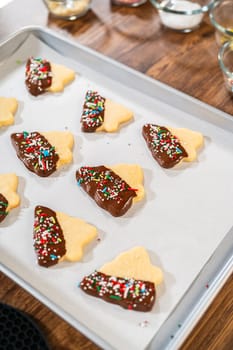 Buttery cookies half-dipped in chocolate, sprinkled with festive red, green, and white decorations, laid on white parchment to set.