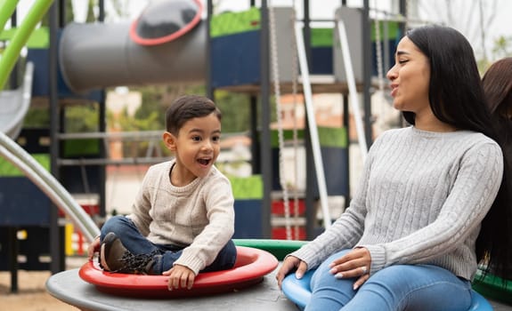 Latin mother and her son are enjoying together on red and blue wheel in a playground as a weekend activity.