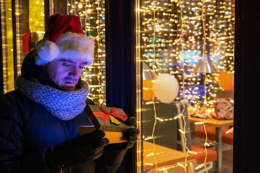 Man in Santas hat with gifts box near illuminated cafe window.