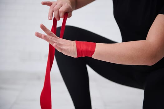 Close-up strong female boxer fighter preparing boxing bandages, wrapping her wrist and hands with a red tape, before wearing boxing gloves, getting ready for training, isolated over white background