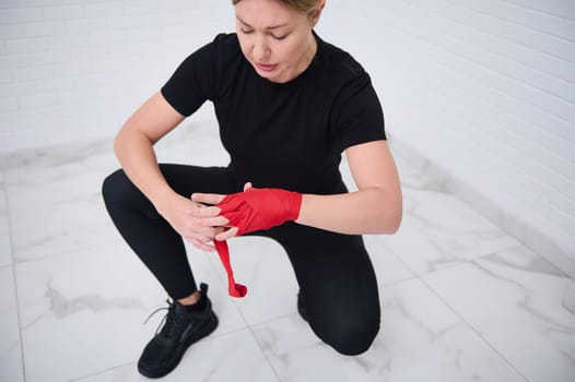 Determined confident European boxer fighter woman 40s, in black sports wear, tying tape around her hand before get boxing gloves preparing to boxing practice. Martial art and combat concept. Top view
