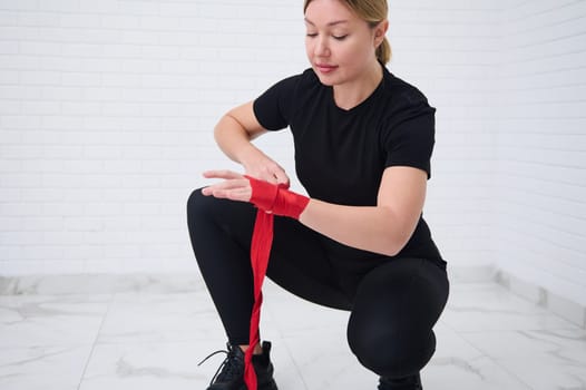 European blonde sporty woman fighter, boxer preparing for boxing competition challenge training, tying tapes before putting boxing gloves, isolated over white background. Sport and martial art concept