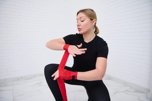 Confident blonde woman boxer fighter preparing for boxing competition challenge training, tying red tapes before putting boxing gloves, isolated over white background. Sport and martial art concept