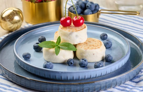 Round small fried cheesecakes on a blue plate with berries. Healthy breakfast, close up