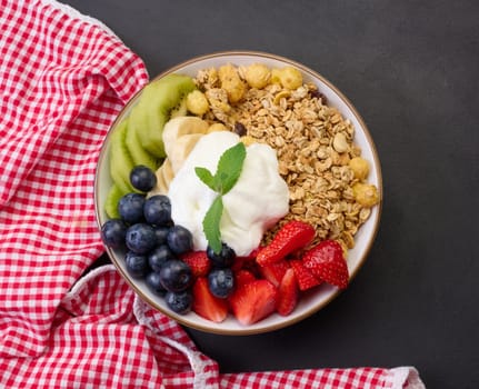 Healthy breakfast. Granola with strawberries, kiwi, banana and blueberries in a round plate on a black table. Healthy and tasty food, top view