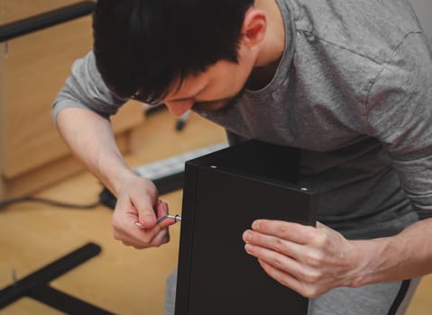 Portrait of one Caucasian young man tightens a screw with a curly wrench connecting a black shelf while sitting on the floor in his room, close-up side view with depth of field. Furniture assembly concept.