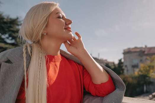 A blonde woman in a red shirt is standing on a balcony. She is smiling and talking on her cell phone