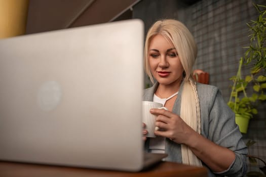 A blonde woman is sitting at a table with a laptop and a white coffee cup. She is wearing a gray jacket and has red lipstick on. The scene suggests a casual and relaxed atmosphere