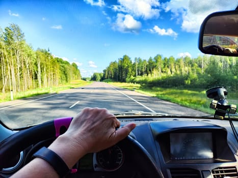 Car salon, steering wheel, hand of woman, view on nature landscape. View from seat of driver on Road, forest, blue sky, white clouds at sunny day. Concept of single trip of female traveller