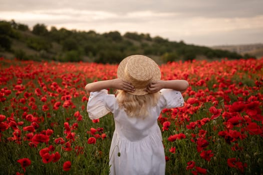 A young girl stands in a field of red poppies, wearing a straw hat and a white dress