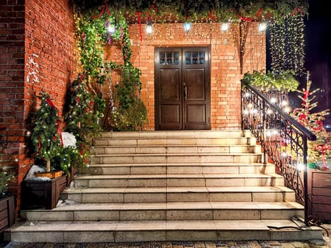 A brightly lit brick porch and staircase adorned with Christmas lights, garlands, and decorated trees at night