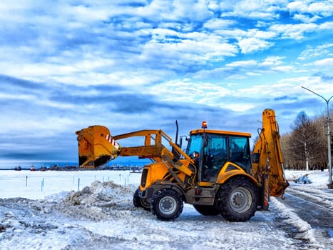 A bright orange tractor is cleaning snow on a winter embankment under a partly cloudy sky, surrounded by snow-covered landscape