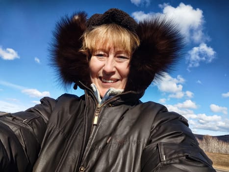 Happy female blonde tourist in a warm jacket taking selfie with outside in cold summer, autumn or winter day. Middle aged woman having fun in nature trip